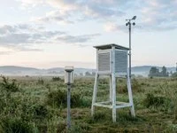 Estação meteorológica ao ar livre com abrigo branco, pluviômetro e anemômetro sob céu de nuvens esparsas