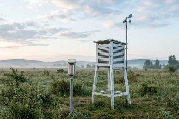 Estação meteorológica ao ar livre com abrigo branco, pluviômetro e anemômetro sob céu de nuvens esparsas